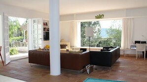 A spacious living room with a brown sofa, a black sofa, and a dining area with a table and chairs. There is a large window with a view of greenery outside. - Superb modern Family Villa nearby fabulous sandy beaches (Rayol-Canadel-Sur-Mer)