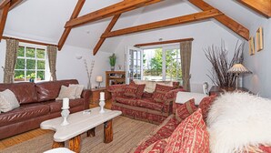 Een gezellige woonkamer met een hoge plafond en zichtbare houten balken, ingericht met een bruine leren bank, een gestreept sofa en een wit, pluizig voetenbankje.