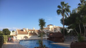 Ein Poolbereich mit einem klaren, blauen Pool, Palmen und einem Gebäude mit einem Kachel­dach im Hintergrund.