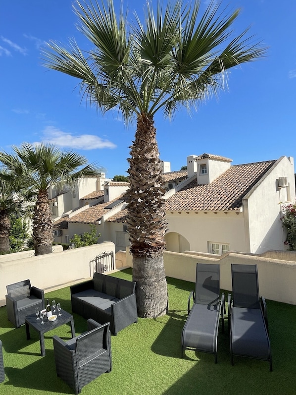 A rooftop patio with outdoor furniture, palm trees, and a view of terracotta-roofed buildings.
