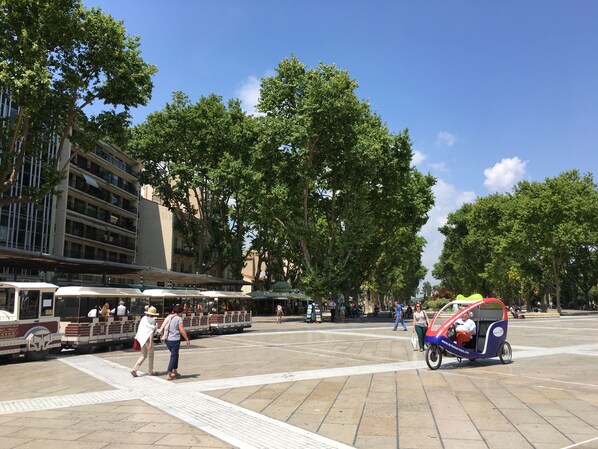 Una plaza pública con un tranvía, personas caminando y un vehículo pequeño.