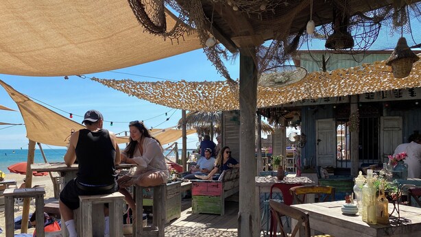 Un espace de restauration en plein air, avec des tables et des chaises en bois, à l’ombre de grands parasols, donnant sur une plage.