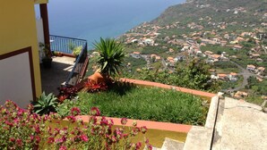 A balcony with potted plants and flowers, overlooking a coastal town with houses and the sea. - Breathtaking Views Holiday House with Heat and or Air conditioning System (Arco da Calheta)