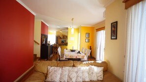 A dining area with a table set for four, a sofa with patterned cushions, and a kitchen in the background.