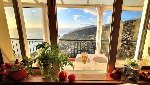 A balcony with a table set for two, a glass of beer, and a view of a coastal town. - Breathtaking Views Holiday House with Heat and or Air conditioning System (Arco da Calheta)