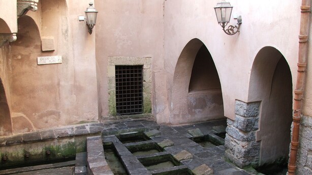 Un patio con escalones de piedra y arcos, una pequeña ventana con celosía metálica y faroles fijados a la pared.