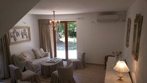 A cozy living room with a sofa, chairs, a coffee table, and a lamp. There is a large window with curtains, an air conditioning unit, and a framed picture on the wall. - Charming house surrounded by vineyards and the Luberon countryside (Grambois)