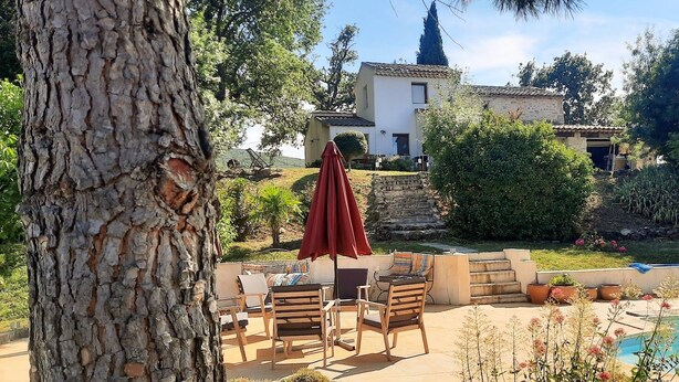 A poolside area with outdoor furniture, a red umbrella, and a view of a house in the background. - Charming house surrounded by vineyards and the Luberon countryside (Grambois)