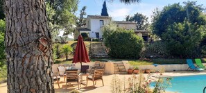 Un espace aménagé au bord de la piscine, avec des meubles d’extérieur, un parasol rouge et une vue sur une maison en arrière-plan.