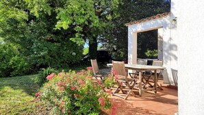 A patio area with a white wall, wooden table, and chairs, surrounded by greenery and trees. - Charming house surrounded by vineyards and the Luberon countryside (Grambois)