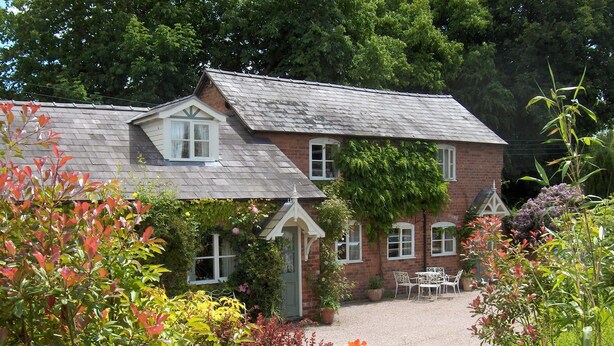 A two-story brick house with a sloped roof, a front porch, and a garden with various plants and flowers.