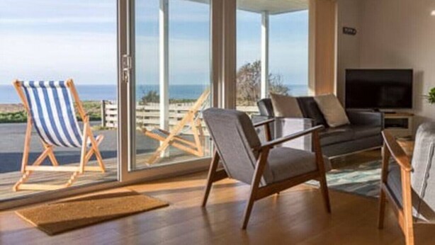A living room with a large TV, a sofa, and two armchairs. There is a sliding glass door leading to an outdoor area with a view of the ocean.