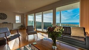 A spacious living room with a large window offering a view of the ocean, a sofa, a coffee table with a vase of flowers, and a clock on the wall.