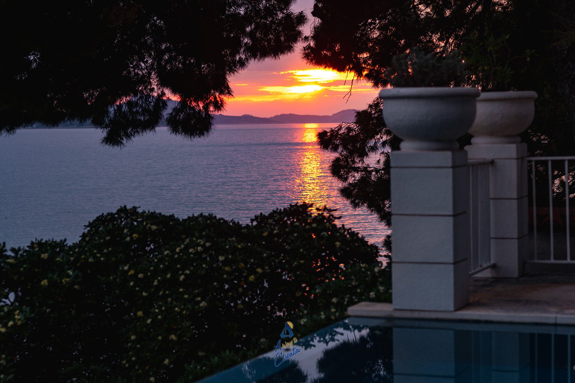 A sunset over a body of water, framed by trees and a building with a pool.