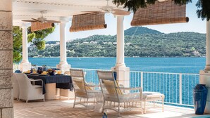 A terrace with wicker chairs, a table set for dining, and a view of a lake and mountains. - Extraordinary seafront villa with pool for up to 9 people (Lozica)