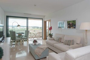 A modern living room with a large window, a dining area with a table and chairs, and a sofa with decorative pillows. - Marina Vilamoura Aquamar 504 by VilamouraSun (Quarteira)