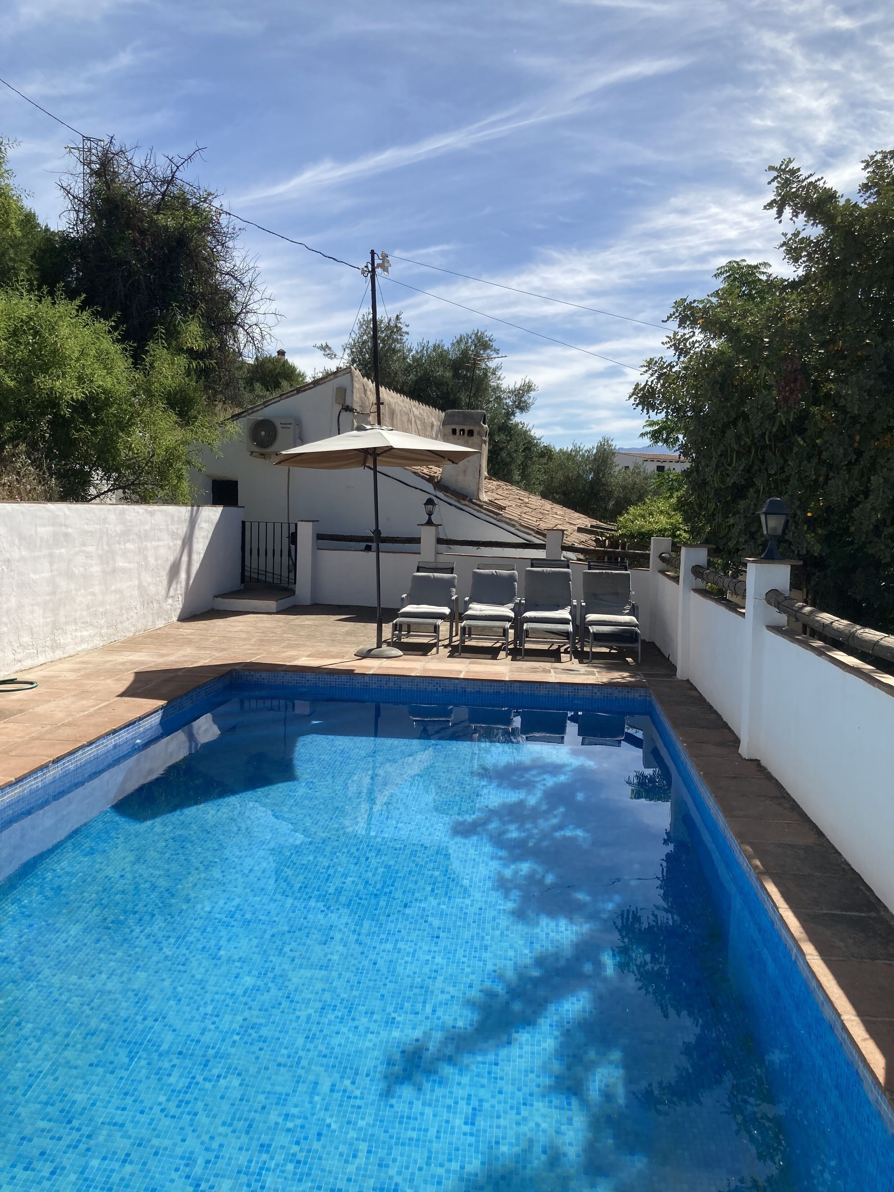 A swimming pool with tiled edges, surrounded by a white wall and a covered patio area with lounge chairs.