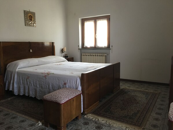A bedroom with a wooden bed, a nightstand, a bedside lamp, a window with curtains, a radiator, and a patterned rug. - Country house "Al Sasso (Città di Castello)