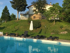 A swimming pool with lounge chairs and an umbrella, surrounded by a grassy hillside and trees. - Country house "Al Sasso (Città di Castello)