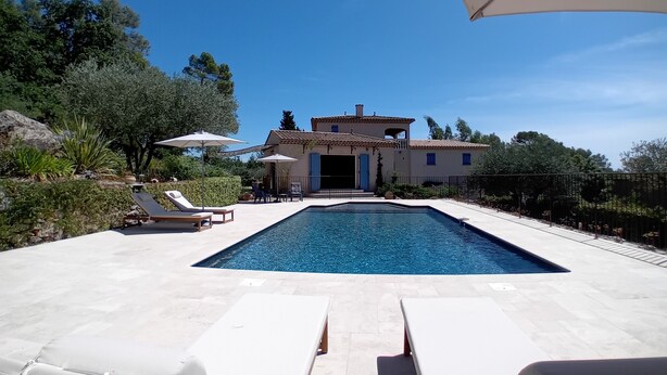 En swimmingpool med solstolar och parasoller, omgiven av en vackert anlagd trädgård och en villa.