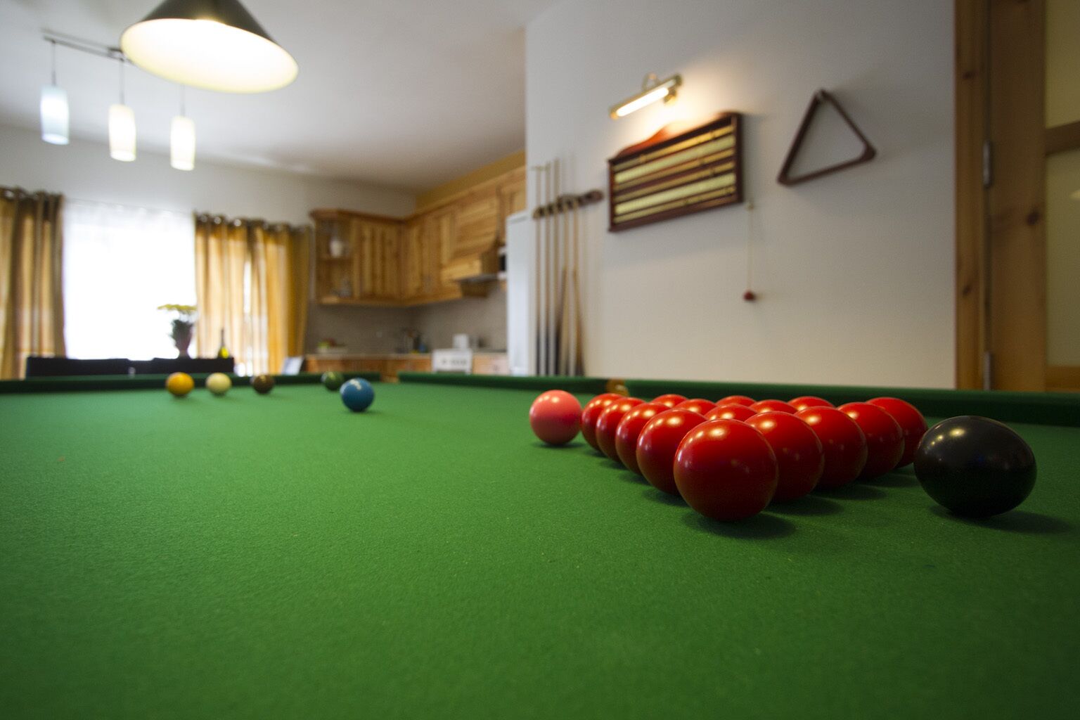 A pool table with red and black balls, a green felt surface, and a kitchen area in the background.
