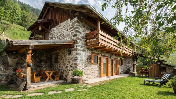A rustic stone house with a wooden balcony, a small outdoor seating area, and a well-maintained lawn. - Cozy Mountain Lodge (Roncegno Terme)