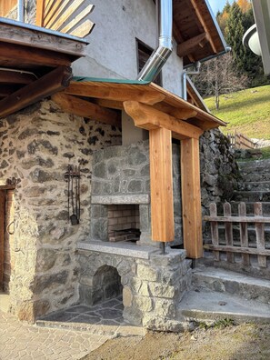 A stone building with a wooden porch, a stone fireplace, and a metal chimney. - Cozy Mountain Lodge (Roncegno Terme)