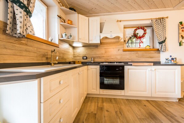 A kitchen with wooden flooring, white cabinetry, a black oven, and a window with a wreath decoration. - Cozy Mountain Lodge (Roncegno Terme)