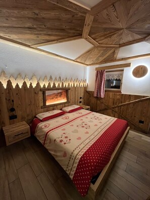 A bedroom with a wooden ceiling, a bed with patterned bedding, a nightstand, and a wall-mounted circular light. - Cozy Mountain Lodge (Roncegno Terme)