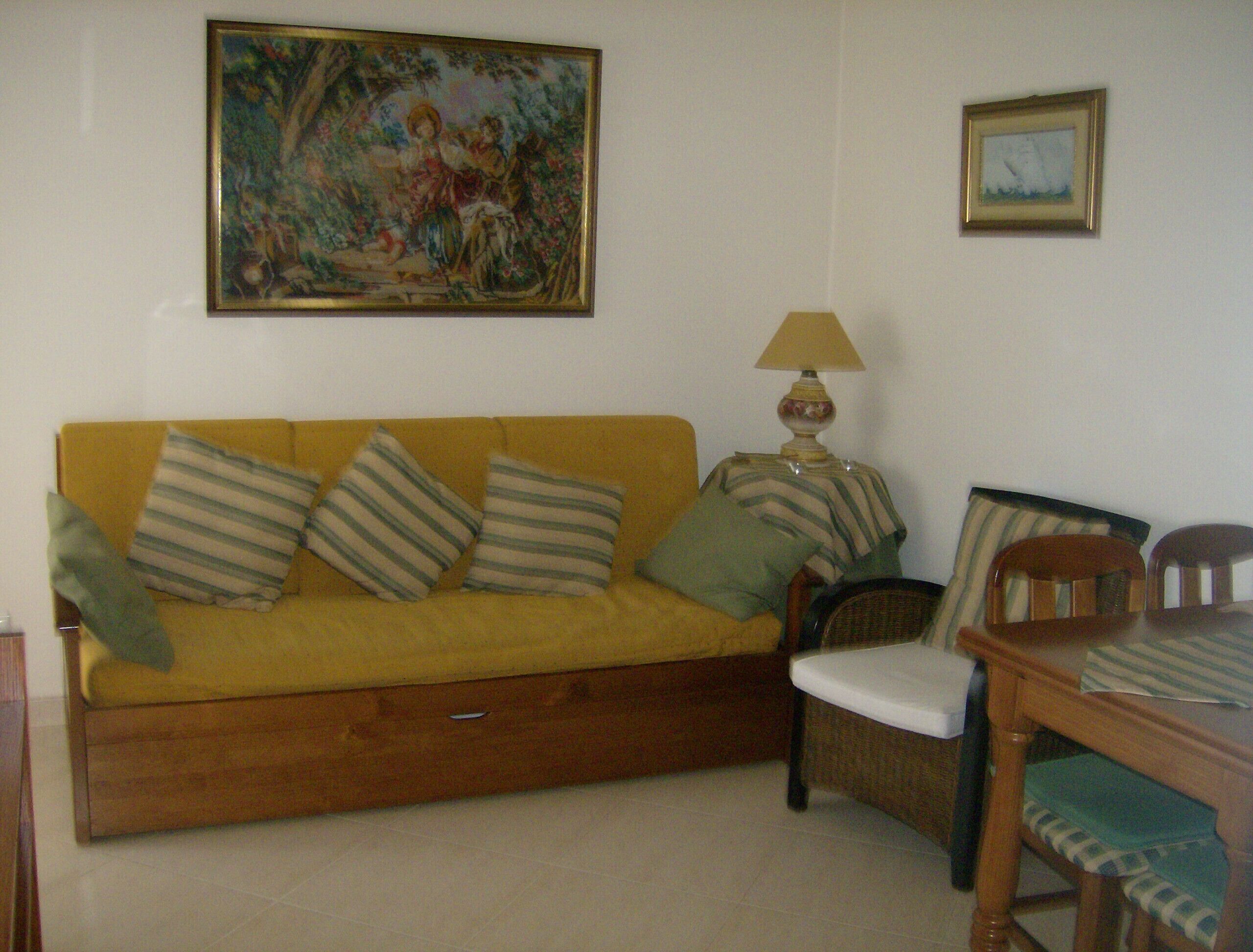 A living room with a yellow sofa, striped pillows, a wooden side table with a lamp, a wicker chair, and two framed pictures on the wall.