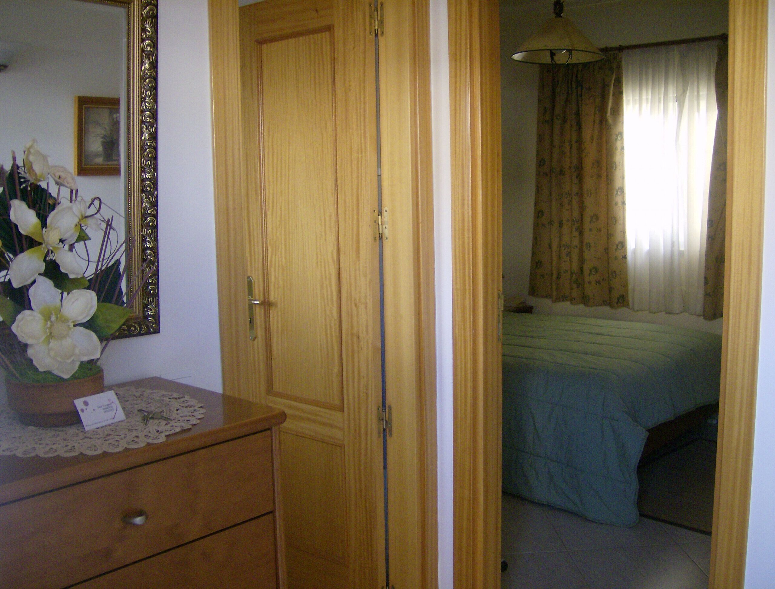 A bedroom with a wooden wardrobe, a bed with a blue bedspread, a nightstand with a vase of flowers, and a mirror on the wall.