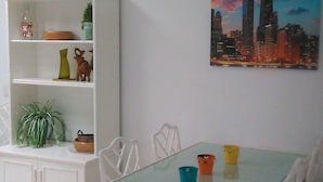 A dining area with a glass table, white chairs, a white cabinet with shelves, and a cityscape painting on the wall. - La Concha Beach. 2Dormitorios wifi and communal pools (Playa Honda)
