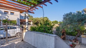 Une cour avec une pergola, une voiture blanche garée dans un espace clos, et un mur de pierre agrémenté de plantes en pot.