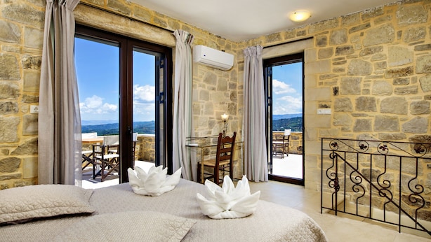 Camera con pareti in pietra, letto con lenzuola bianche, balcone con tavolo e sedie e vista sul paesaggio.