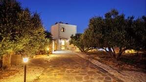 Un sentiero in pietra conduce a un edificio con una piccola finestra e una porta, circondato da alberi e un'area ben illuminata.