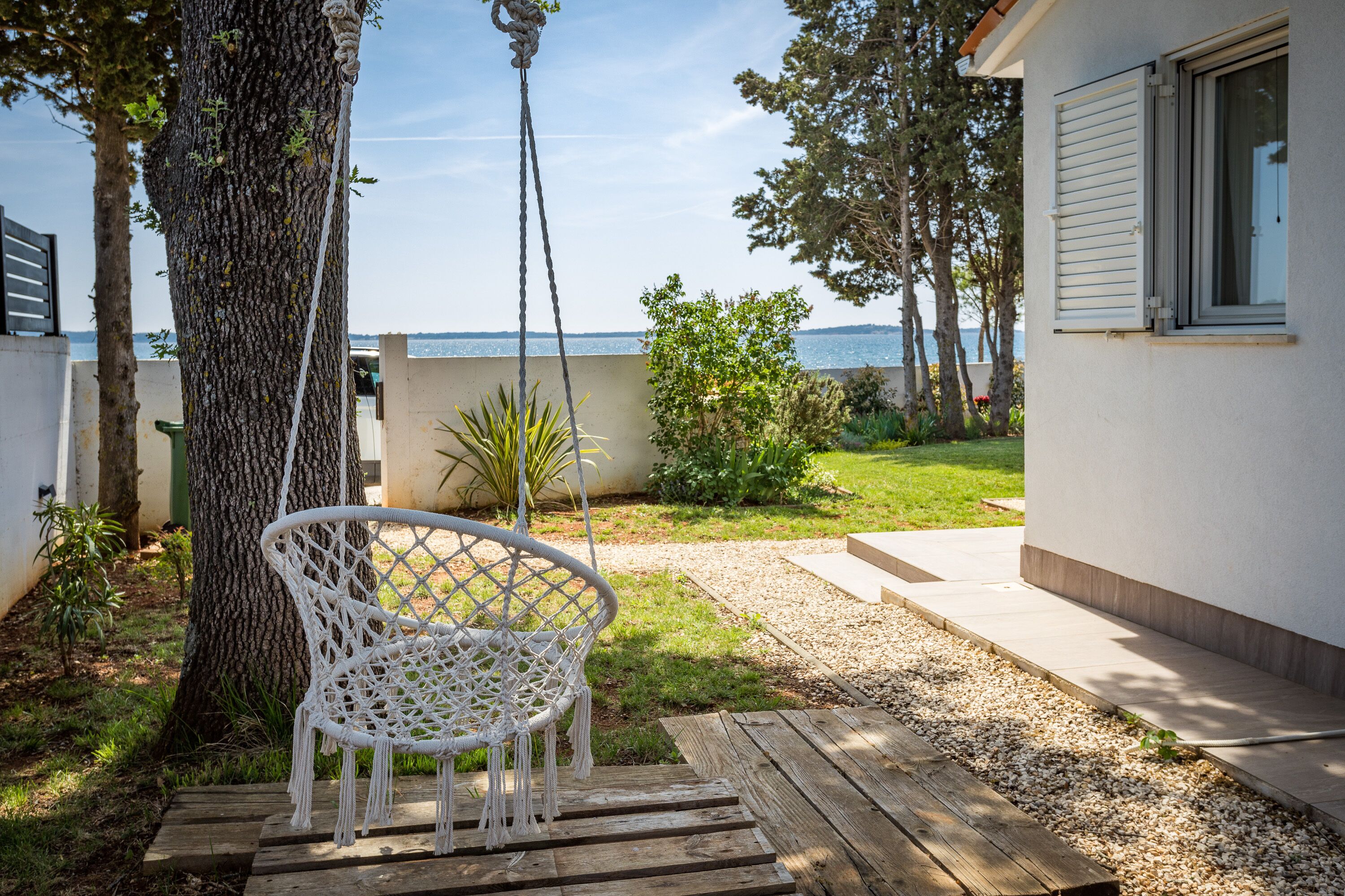 A white wicker chair hanging from a tree, a wooden deck, and a view of the sea.