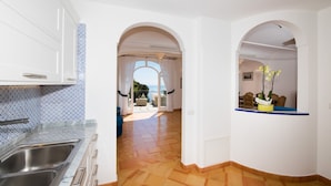 Eine Küche mit eingebautem Spülbecken, weißen Schränken und einer ornamentierten Spritzschutzwand. Ein Gang führt zu einem Balkon mit Blick aufs Meer.