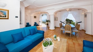 Ein geräumiges Wohnzimmer mit einem blauen Sofa, einer Essecke mit Tisch und Stühlen und Meerblick durch einen offenen Durchgang.