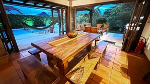 Une table à manger en bois, sur laquelle se trouve un panier de fruits, entourée de chaises, dans un espace extérieur abrité offrant une vue sur un jardin et un hamac.