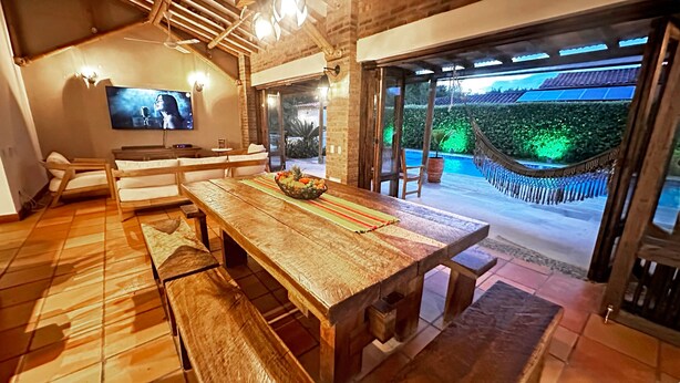 Une table à manger en bois avec des bancs, un téléviseur fixé au mur et une vue sur une piscine avec un hamac.