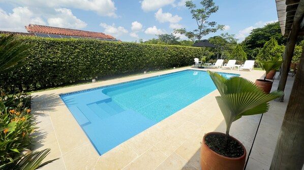 Une piscine rectangulaire dont le bord est recouvert de carreaux, entourée d’une haie basse et de plantes en pot.