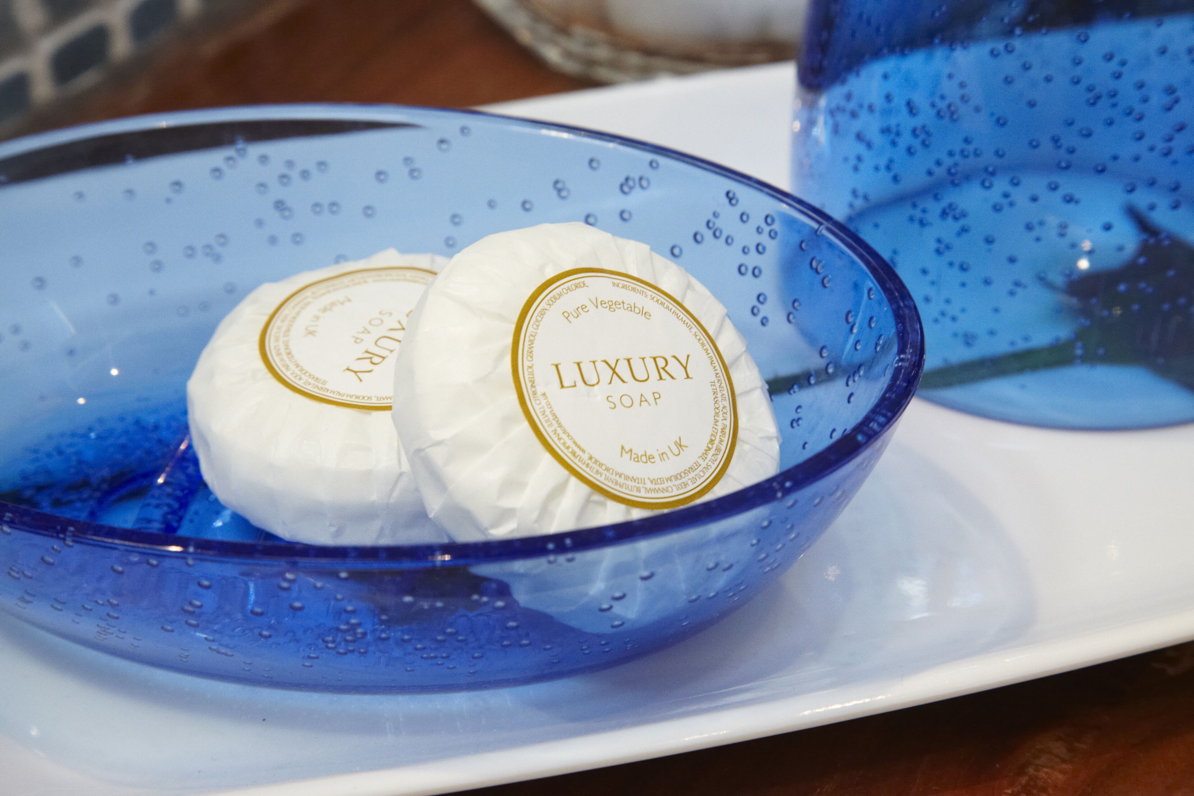 Un tazón de vidrio azul contiene dos pastillas de jabón blanco con la etiqueta 'Luxury Soap' sobre un plato blanco.
