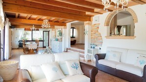 A spacious living area with a wooden ceiling, a white sofa with decorative pillows, a brown leather sofa, and a chandelier.
