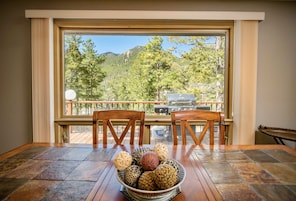 Comedor con un ventanal grande que da a una terraza y montañas. Hay una mesa con sillas y un cuenco de objetos decorativos.