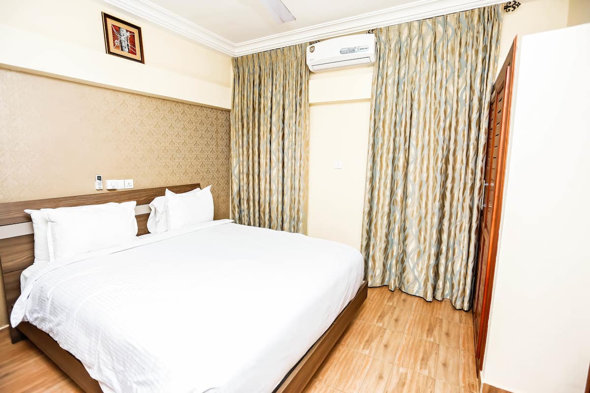 A hotel room with a large bed, wooden headboard, beige walls, and a patterned curtain.