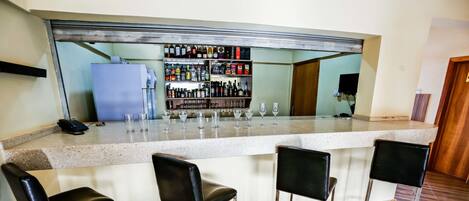 A bar area with a counter, three black bar stools, and a shelf with various bottles and glasses.