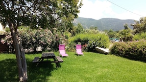 A grassy outdoor area with a picnic table and two pink lounge chairs, surrounded by lush greenery and flowering bushes, with a mountain view in the background.
