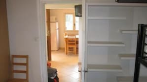A room with a wooden chair, a white wardrobe, and a kitchen visible through an open door.