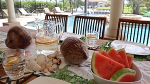A table set with a pitcher, glasses, coconuts, and watermelon slices, with a pool and lounge chairs in the background. - Villagoona, waterfront, infinity pool, dir.access to the Gulf of America (Cape Coral)