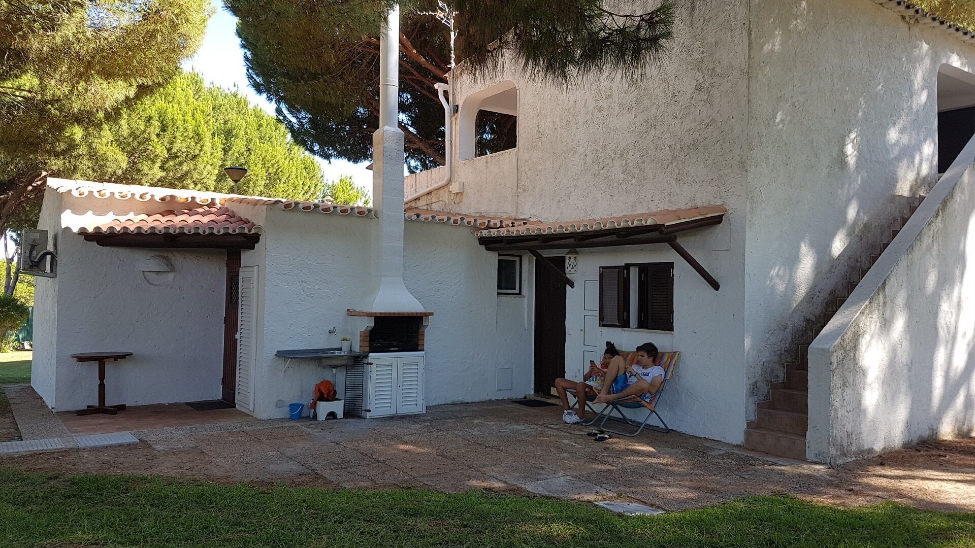 A rustic outdoor kitchen area with a chimney, a small table, and a seating area with a person relaxing.